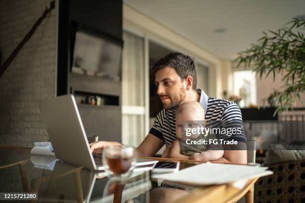 hombre sosteniendo a su hijo recién nacido y trabajando con computadora portátil en casa - banca electrónica fotografías e imágenes de stock
