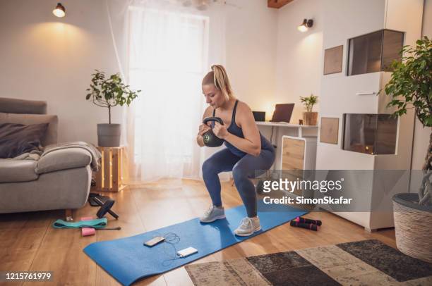mujer joven en cuclillas con kettlebell en estera de yoga en la sala de estar - crouching fotografías e imágenes de stock