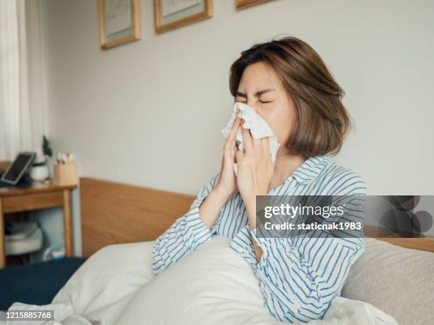 asiatische frau bekam grippe niesen in gewebe sitzen auf dem bett - sinusitis stock-fotos und bilder