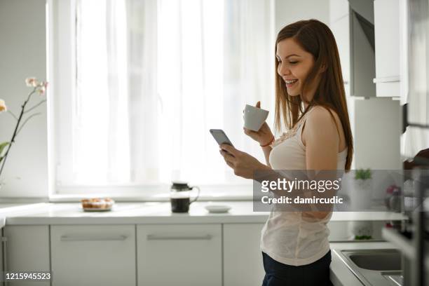 jonge vrouw die koffie drinkt en slimme telefoon thuis gebruikt - woman drinking phone kitchen stockfoto's en -beelden