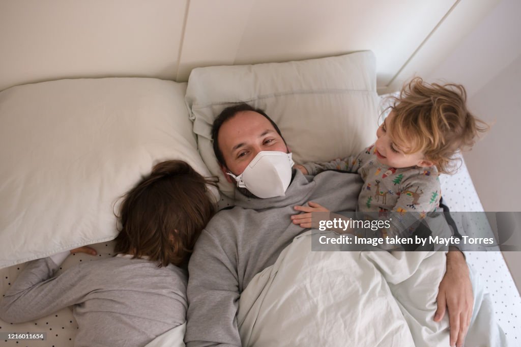 Dad wearing surgical mask lying in bed with sons