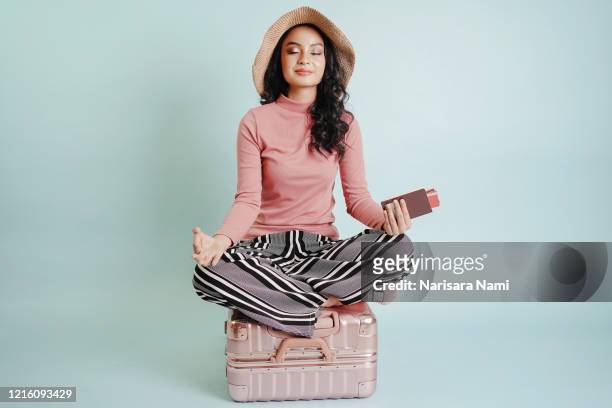 happy tourist sitting on the suitcase, pretending yoga on a suitcase and hand holding passport and credit card isolated on blue background with copy space. dreams about traveling concept. - indonesian girl stock pictures, royalty-free photos & images