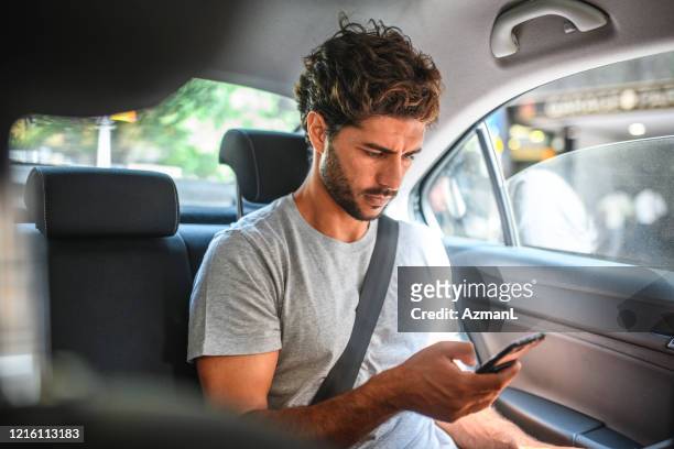 joven hispano macho que comprueba el teléfono inteligente en el asiento trasero del coche - passenger fotografías e imágenes de stock