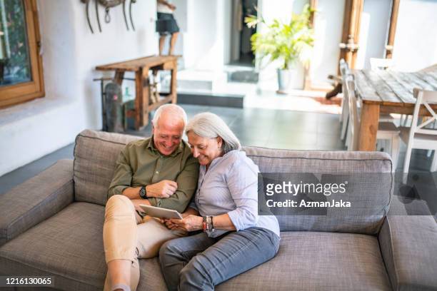 senior couple sitting together on sofa with digital tablet - two houses side by side stock pictures, royalty-free photos & images