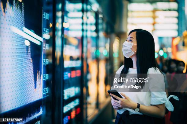 economic and financial impact during the covid-19 health crisis deepens. businesswoman with protective face mask checking financial trading data on smartphone by the stock exchange market display screen board in downtown financial district showing stock m - economia foto e immagini stock