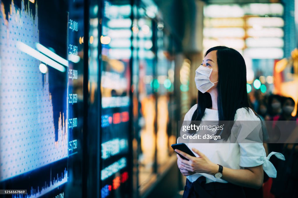 Economic and financial impact during the Covid-19 health crisis deepens. Businesswoman with protective face mask checking financial trading data on smartphone by the stock exchange market display screen board in downtown financial district showing stock m