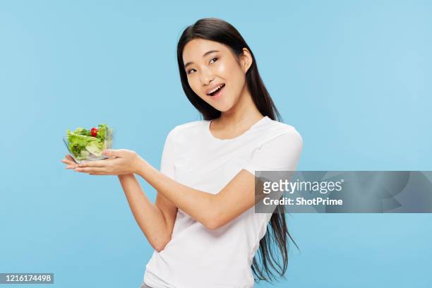 young asian beautiful woman in a white t-shirt on a blue background holds in her hands a cup with salad and vegetarian food. - national diet of japan stock pictures, royalty-free photos & images