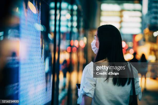 economic and financial impact during the covid-19 health crisis deepens. businesswoman with protective face mask checking financial trading data on smartphone by the stock exchange market display screen board in downtown financial district showing stock m - global business covid stock pictures, royalty-free photos & images