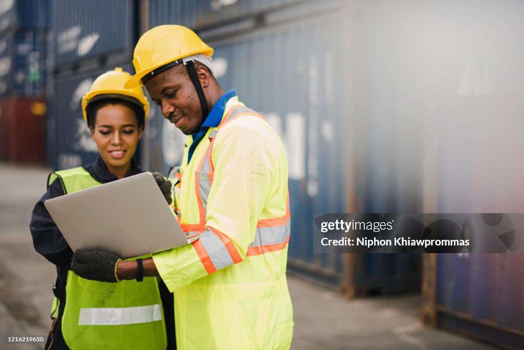 Young African american men and woman worker Check and control loading freight Containers by use computer laptop at commercial shipping dock felling happy. Cargo freight ship import export concept