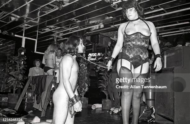 An unidentified dominatrix, dressed in lingerie and stockings, hold the leash of a naked man in wrist restraints who kneels on the floor during an...