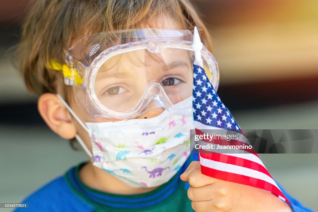 American little caucasian boy posing with a protective mask, protective glasses holding a US flag