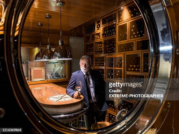 elderly rich man savoring white wine on the other side of the porthole in a wine cellar of a yacht - wine cellar stock pictures, royalty-free photos & images
