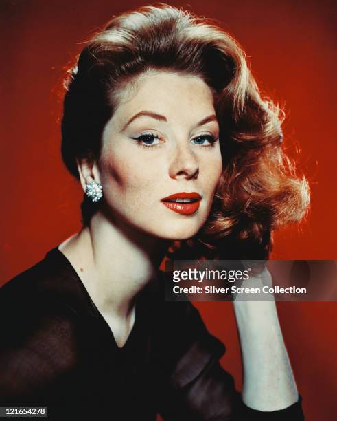 Suzy Parker , US actress and model, wearing a black chiffon top and diamond earrings in a studio portrait, against a red beckground, circa 1955.