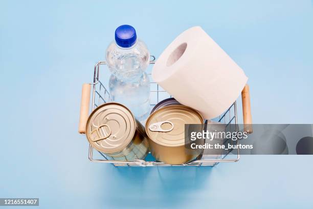 still life of canned food, bottle of mineral water and toilet paper in shopping basket on blue background - buying toilet paper stock pictures, royalty-free photos & images