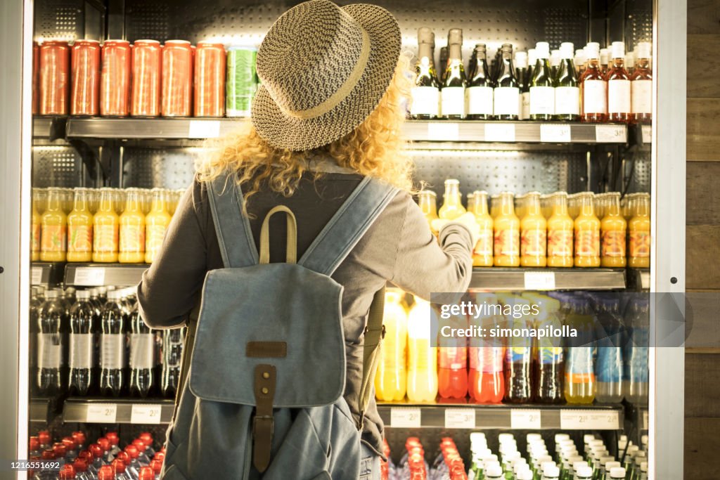 Travel woman viewed from back choosinf beverage in a fresh fridge - airport or station bar concept and traveler passenger choosing drinks - modern lifestyle backpack people buying drinks bottles