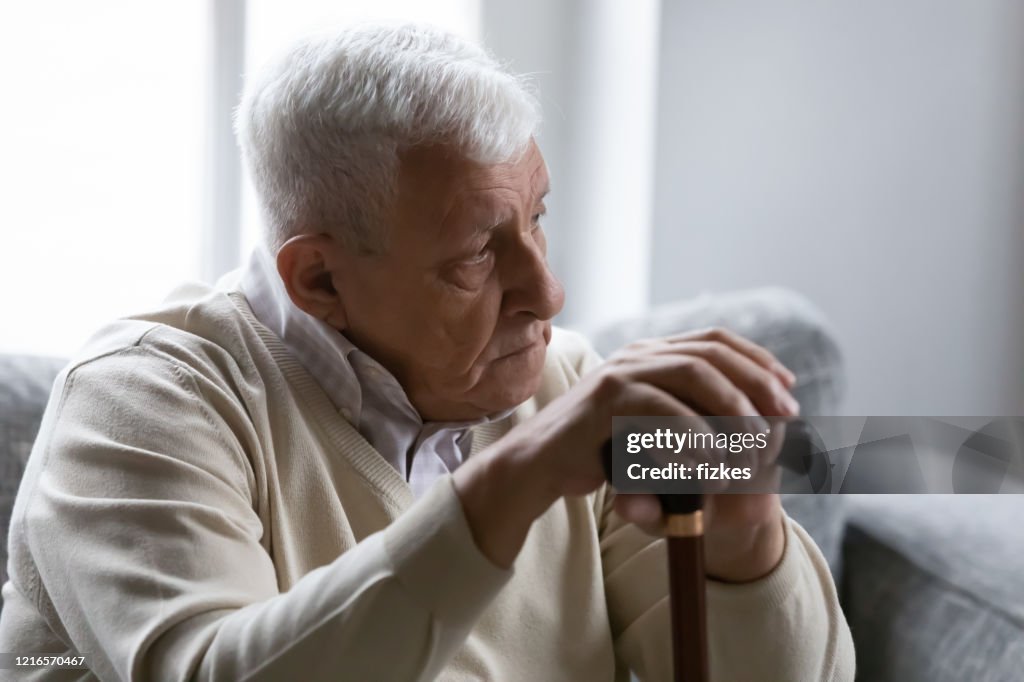 Old man holds cane sitting on couch looking at distance