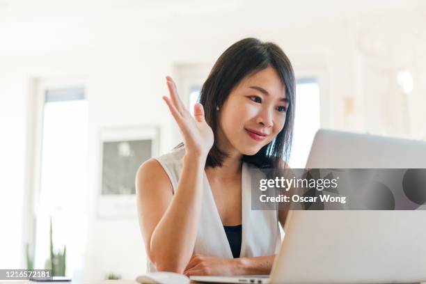 cheerful young woman waving at laptop during a video conference at home - zwaaien gebaren stockfoto's en -beelden