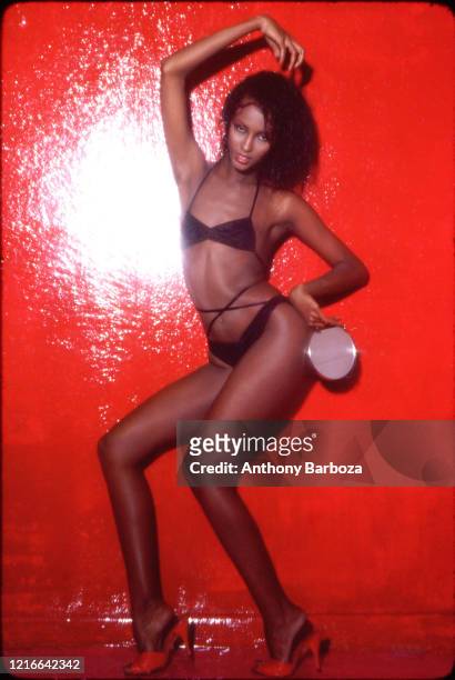 Portrait of Somali-born American fashion model Iman, dressed in a black bikini and heels, as she poses against a red background, late 1970s or early...