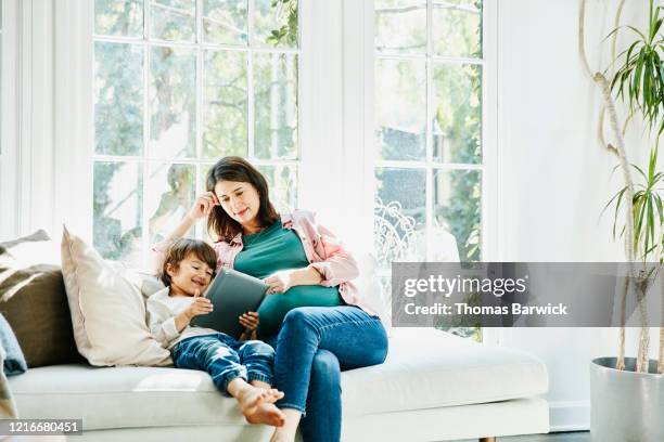 pregnant mother and young son watching video on digital tablet while sitting on couch in living room - pregnancy class stockfoto's en -beelden