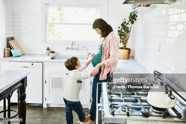 Smiling young son putting hand on pregnant mothers stomach while standing in home kitchen
