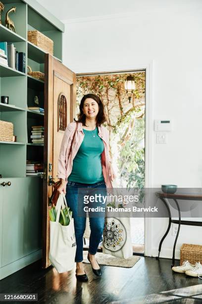 smiling pregnant woman carrying groceries in canvas bags into home - entering photos et images de collection