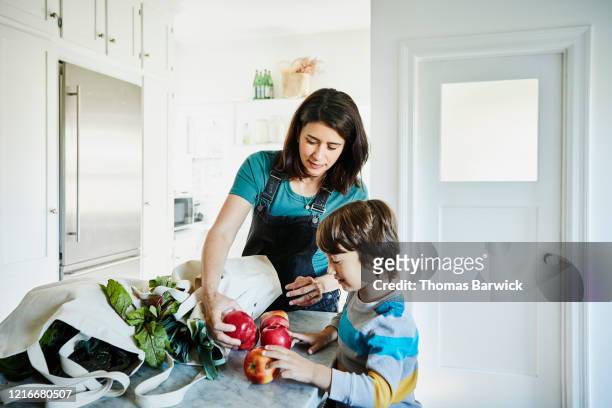 pregnant mother showing young son apples in kitchen after shopping for groceries - child holding apples stockfoto's en -beelden