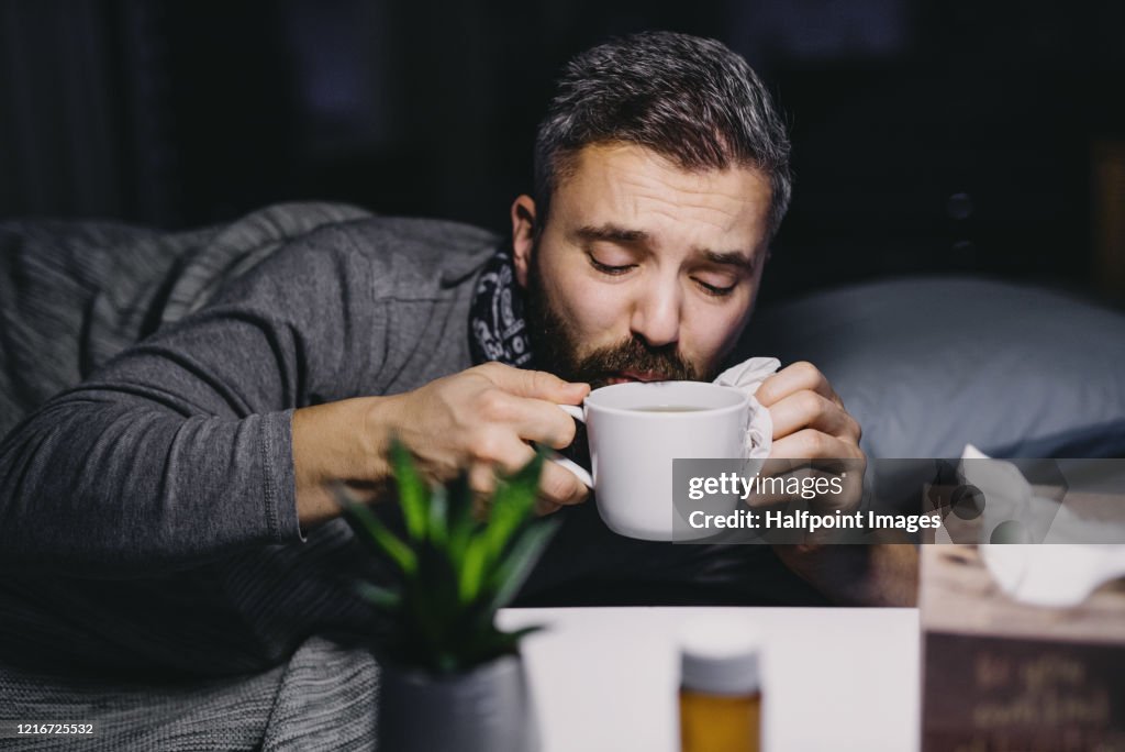 An ill mature man patient in bed at home, drinking hot tea.