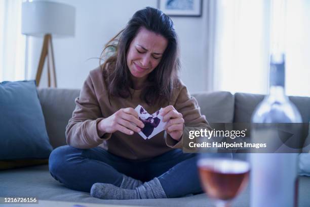 sad and depressed woman sitting indoors on sofa, tearing photograph. - breaking up stock pictures, royalty-free photos & images