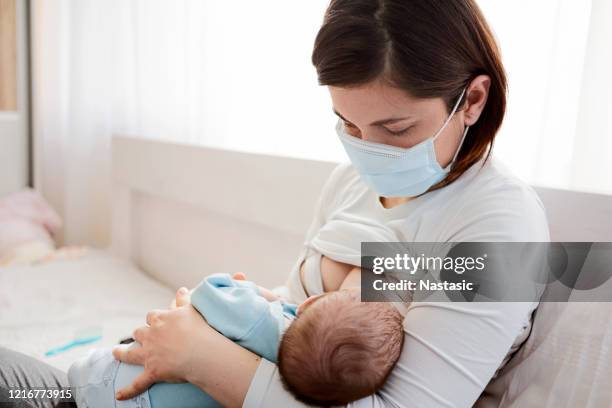 mother with protective mask breastfeeding her baby sitting on a bed - serbia covid stock pictures, royalty-free photos & images