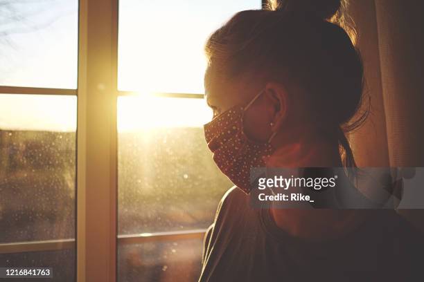 pretty woman in quarantine with mask, looks out the window - lockdown stock pictures, royalty-free photos & images