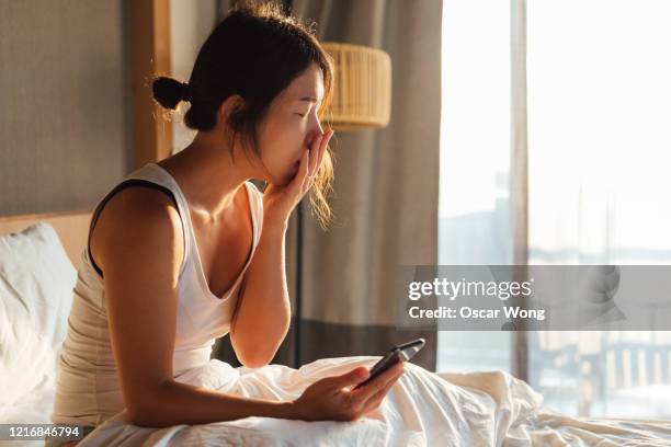 young woman feeling tired using mobile phone in bed - yawning is contagious stock-fotos und bilder