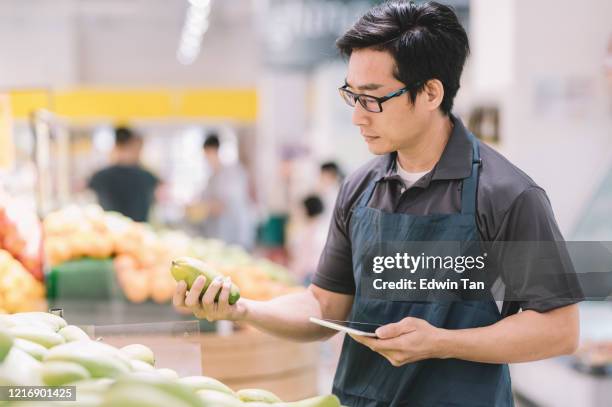 an asian chinese male store assistant checking inventory and price tag in front of fruit store department with his digital tablet - digital price tag stock pictures, royalty-free photos & images