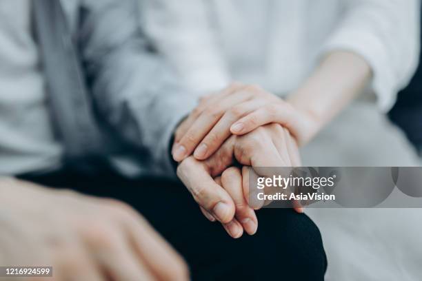 colpo ritagliato di una giovane coppia seduta sul divano che si tiene compassionevolmente per mano in autoisolamento durante la crisi sanitaria covid-19 - incoraggiamento foto e immagini stock