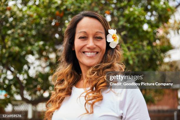 reife frau steht draußen mit einer blume in den haaren und lächelnd - hawaiianischer-abstammung stock-fotos und bilder