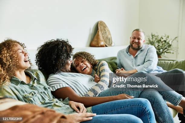 laughing daughter embracing mother while sitting on couch with family in living room - young couple photos et images de collection