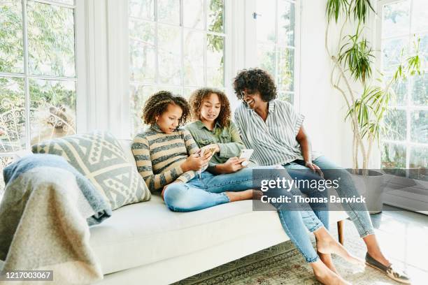 smiling mother and daughters sitting on couch in living room looking at smartphone - girls barefoot in jeans fotografías e imágenes de stock