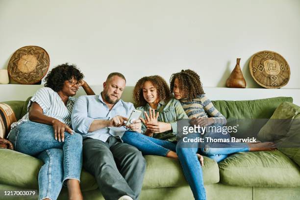 family sitting on couch in living room looking at smart phone - four people stock pictures, royalty-free photos & images