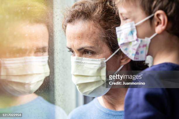 mulher madura posando com seu filho, ambos usando máscaras de proteção, muito triste olhando pela janela preocupado com o bloqueio covid-19 - stay home - fotografias e filmes do acervo