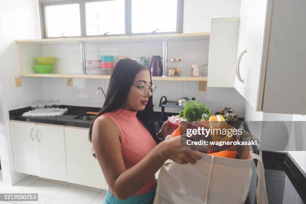 joven desempaquetando comestibles de su bolsa de compras reutilizable en la cocina - frugalidad fotografías e imágenes de stock