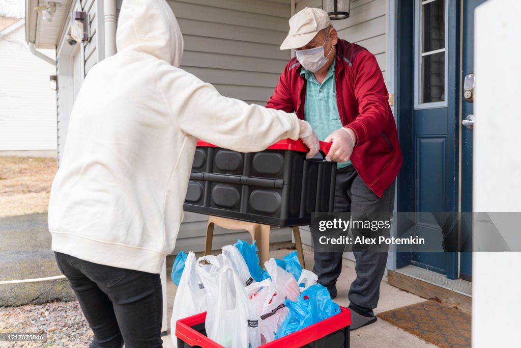 Young woman wearing a protective n95 mask, a courier, is handing the groceries packed into reusable plastic bins to a customer, a senior man, on the porch of his house during COVID-19 outbreak.