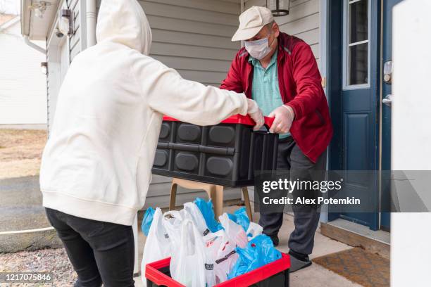 young woman wearing a protective n95 mask, a courier, is handing the groceries packed into reusable plastic bins to a customer, a senior man, on the porch of his house during covid-19 outbreak. - serviços-essenciais imagens e fotografias de stock