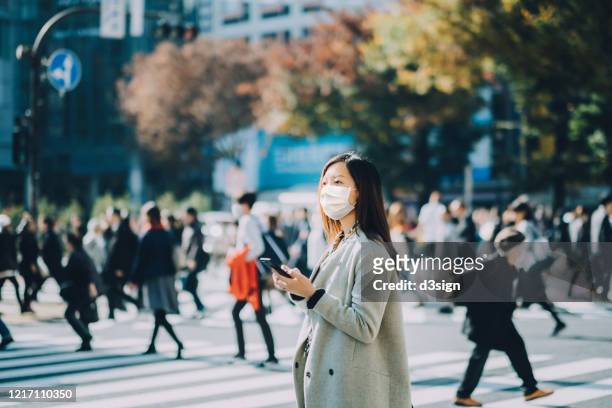 young asian businesswoman commuting in busy downtown city street with protective face mask and using smartphone to protect and prevent from the spread of viruses during covid-19 health crisis - 通勤 スト��ックフォトと画像