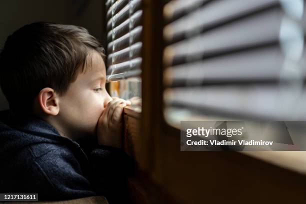 small sad boy looking through the window during coronavirus isolation. - loneliness stock pictures, royalty-free photos & images