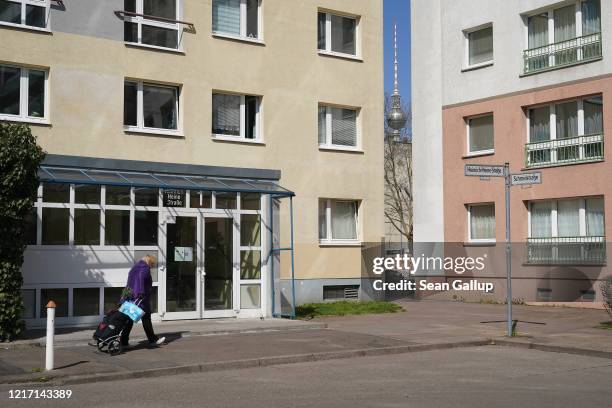 An elderly woman pulling a shopping cart laden with toilet paper walks among residential apartment buildings as the broadcast tower at Alexanderplatz...