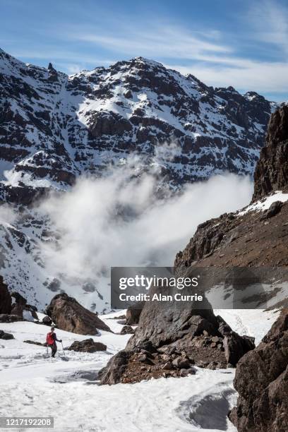 hiker descending in snow into a valley at toubkal mountain, morocco. on a sunny day with clouds in the valley below and rocky landscape - atlas mountains stock pictures, royalty-free photos & images