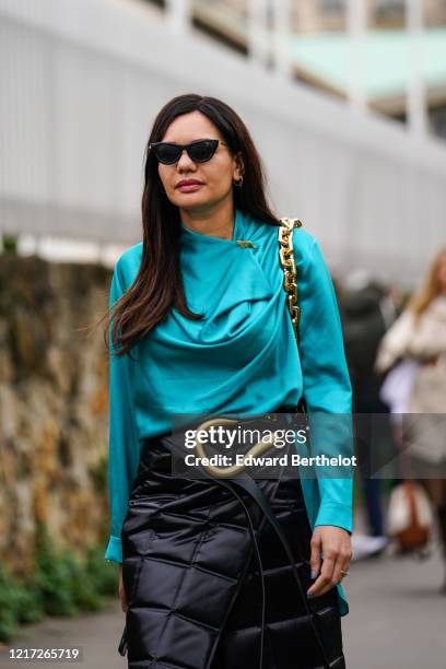 Guest wears sunglasses, earrings, a turquoise-blue draped top, a black quilted wrap-over skirt, black tights, a black belt with a large gold-tone...