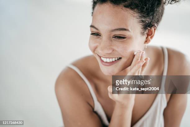 smiling young women applying moisturiser to her face - skin care face stock pictures, royalty-free photos & images