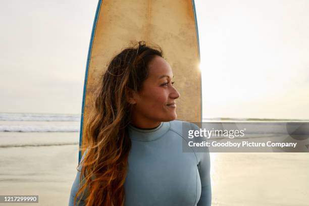 lächelnde reife frau steht an einem strand mit ihrem surfbrett - beach hold surfboard stock-fotos und bilder