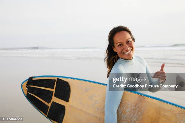 lächelnde frau zu fuß mit ihrem surfbrett und werfen das shaka-zeichen - shaka sign stock-fotos und bilder