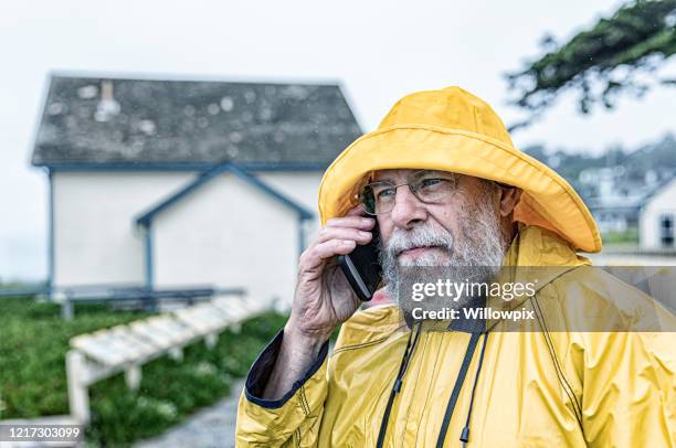 senior man wearing yellow raincoat using mobile phone - rain hat stock pictures, royalty-free photos & images
